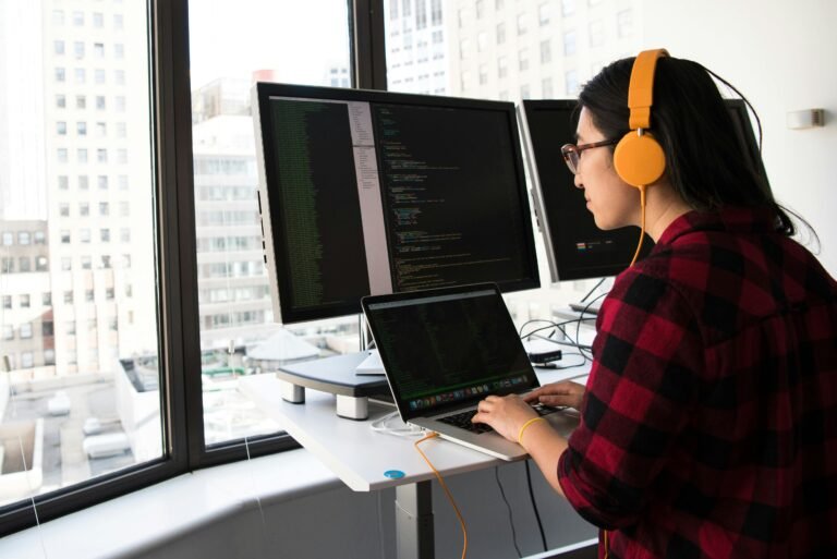 Woman programming on a laptop at a standing desk in an office with large windows.