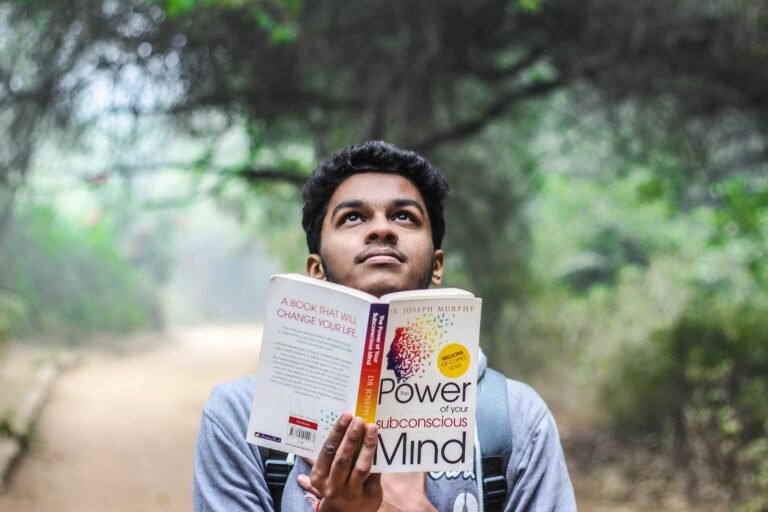 A young man relaxes while reading 'Power of Your Subconscious Mind' outdoors in a park.